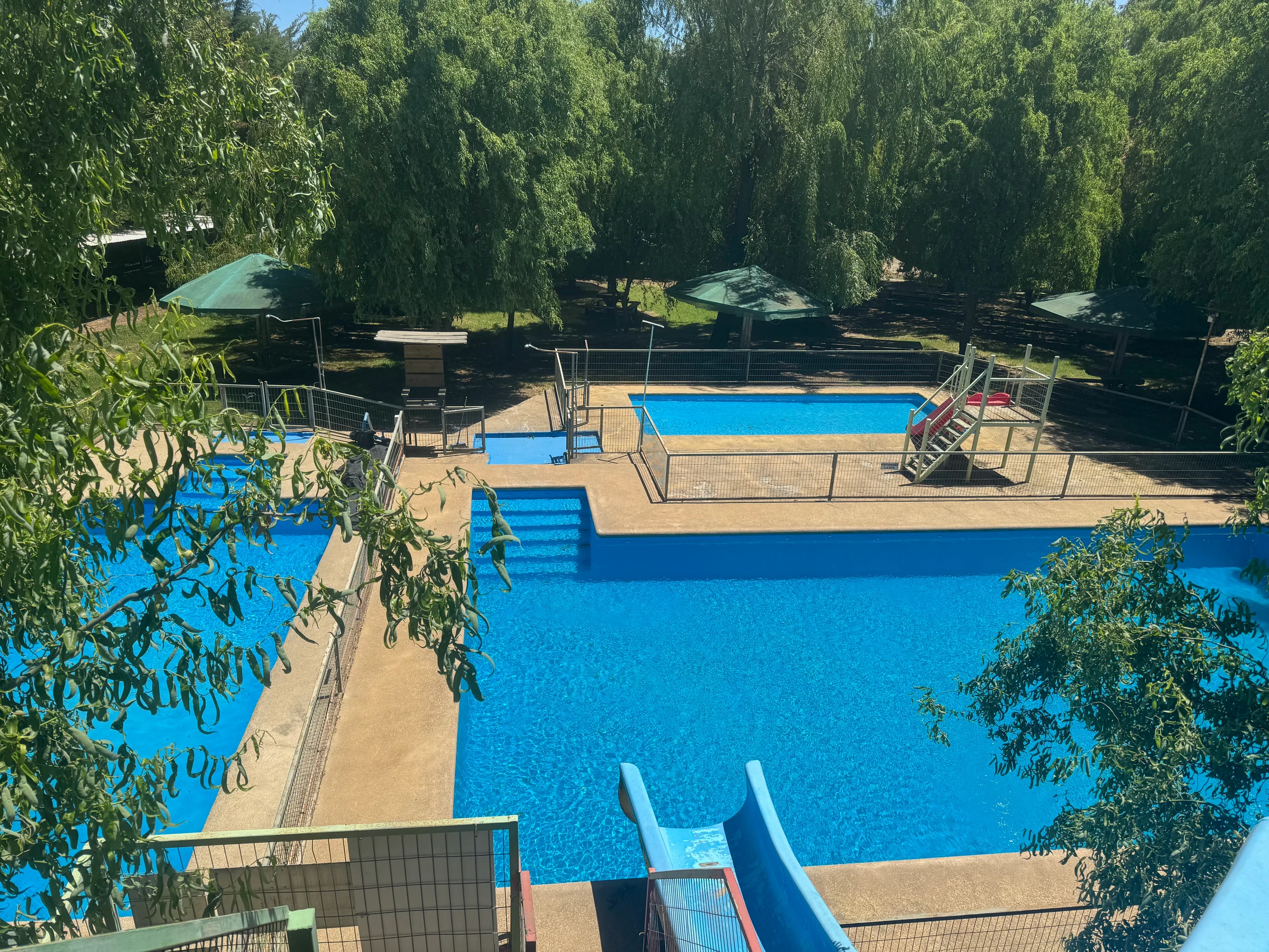 Vista elevada de las piscinas del sector 1, con tobogán azul y áreas de descanso bajo los sauces